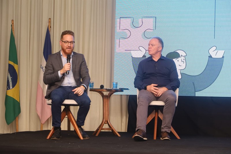 Dois homens brancos, em trajes sociais de tom cinza, estão sentados a frente de uma tela enquanto discursam com microfones.
